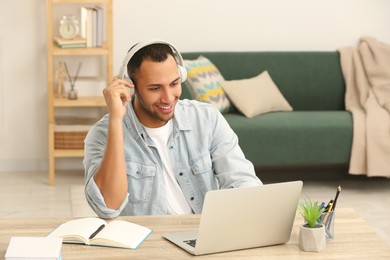 Photo of African American man in headphones working on laptop at wooden table in room
