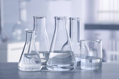 Photo of Different laboratory glassware with transparent liquid on wooden table against blurred background