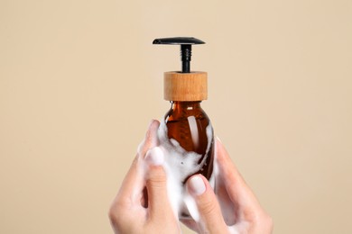 Photo of Woman with foam on hands holding bottle of skin cleanser against beige background, closeup. Cosmetic product