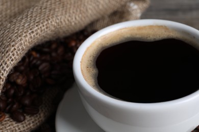 Cup of hot aromatic coffee and sack with roasted beans, closeup