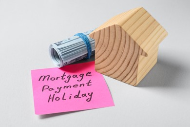 Photo of Paper note with words Mortgage Payment Holiday, money and house model on light grey background, closeup