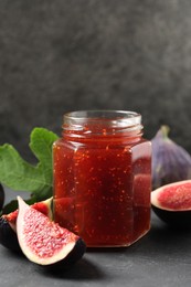 Glass jar with tasty sweet jam, green leaf and fresh figs on grey table, closeup