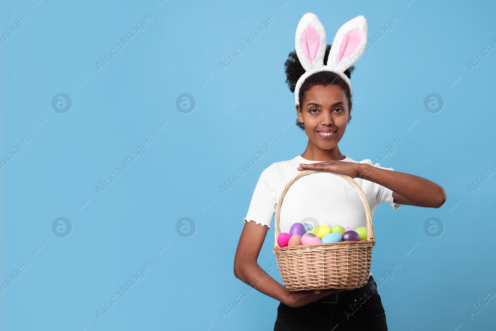 Photo of Happy African American woman in bunny ears headband holding wicker basket with Easter eggs on light blue background, space for text