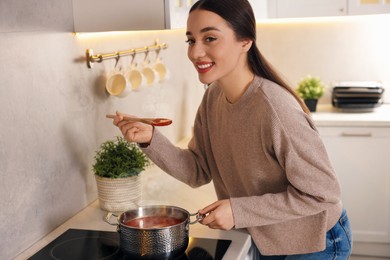 Smiling woman with wooden spoon tasting tomato soup in kitchen