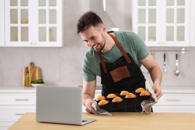 Photo of Man holding muffins near laptop at table in kitchen. Time for hobby