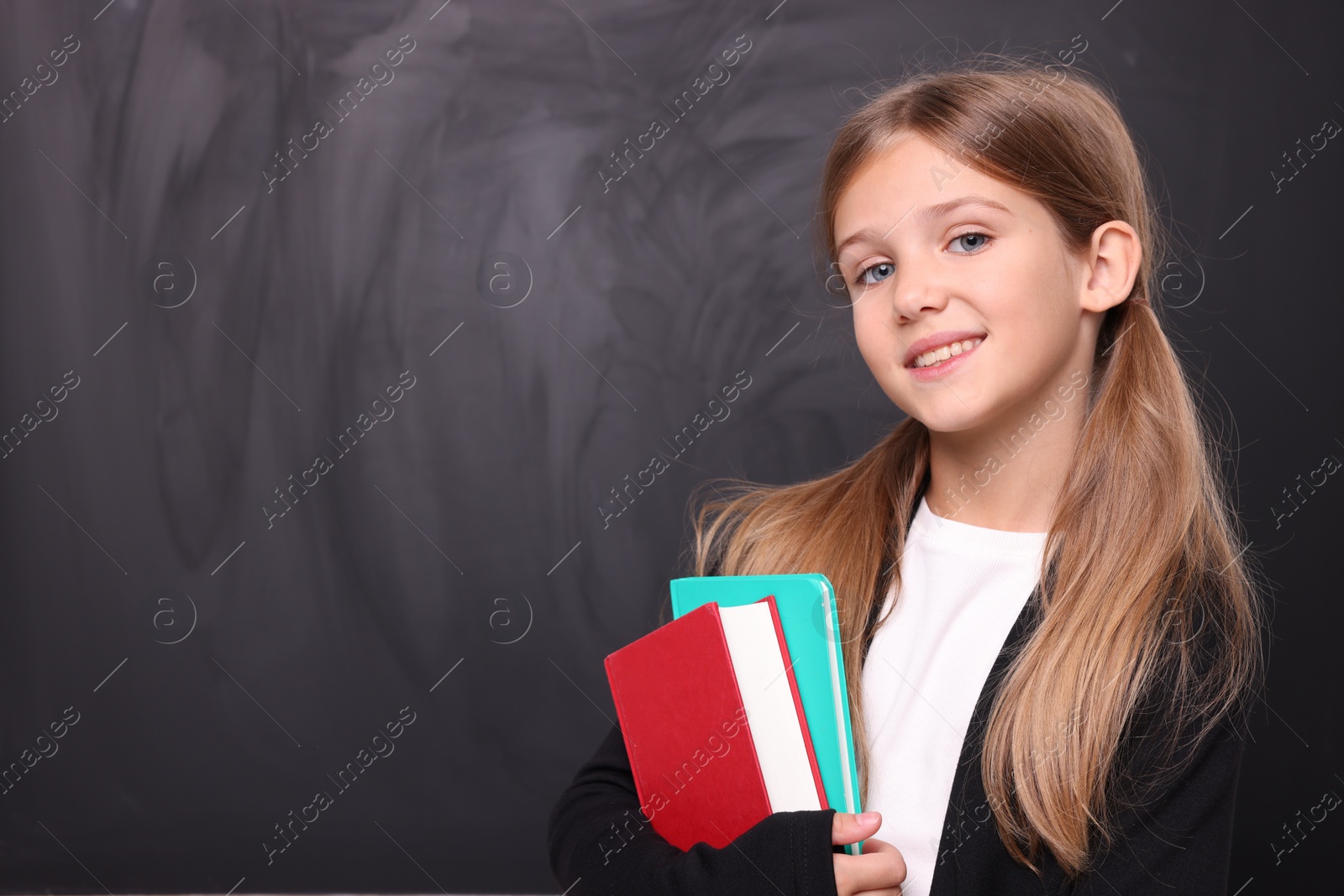 Photo of Smiling schoolgirl with books near blackboard. Space for text