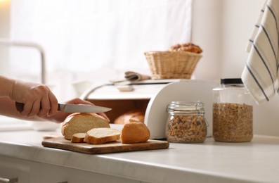 Woman cutting loaf near wooden bread basket at white marble table in kitchen, closeup