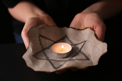 Photo of Woman holding fabric with star of David and burning candle on black background, closeup. Holocaust memory day