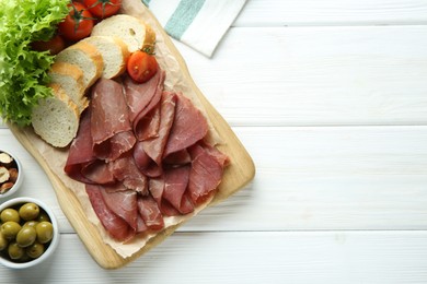 Photo of Board with delicious bresaola served with tomato, bread and lettuce on white wooden table, flat lay. Space for text