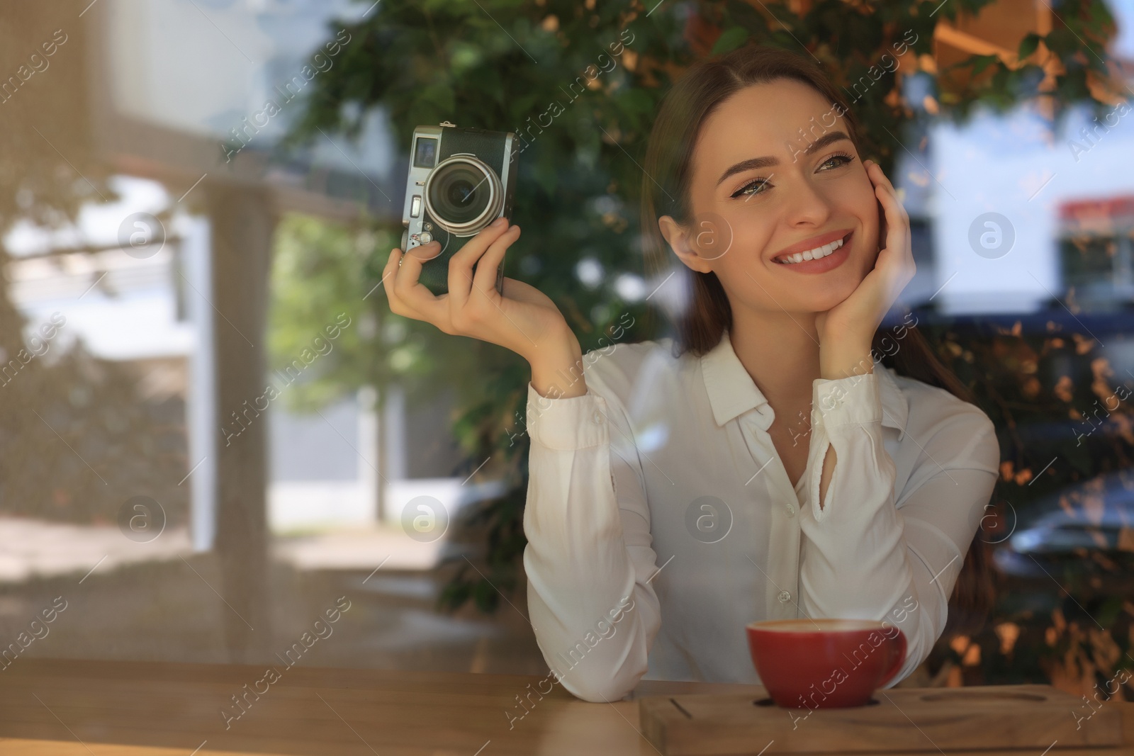 Photo of Young woman with camera and cup of coffee at cafe, view through window. Creative hobby