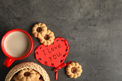 Photo of Heart shaped greeting card with phrase I Love You near cookies and red cup on dark grey table, flat lay. Space for text