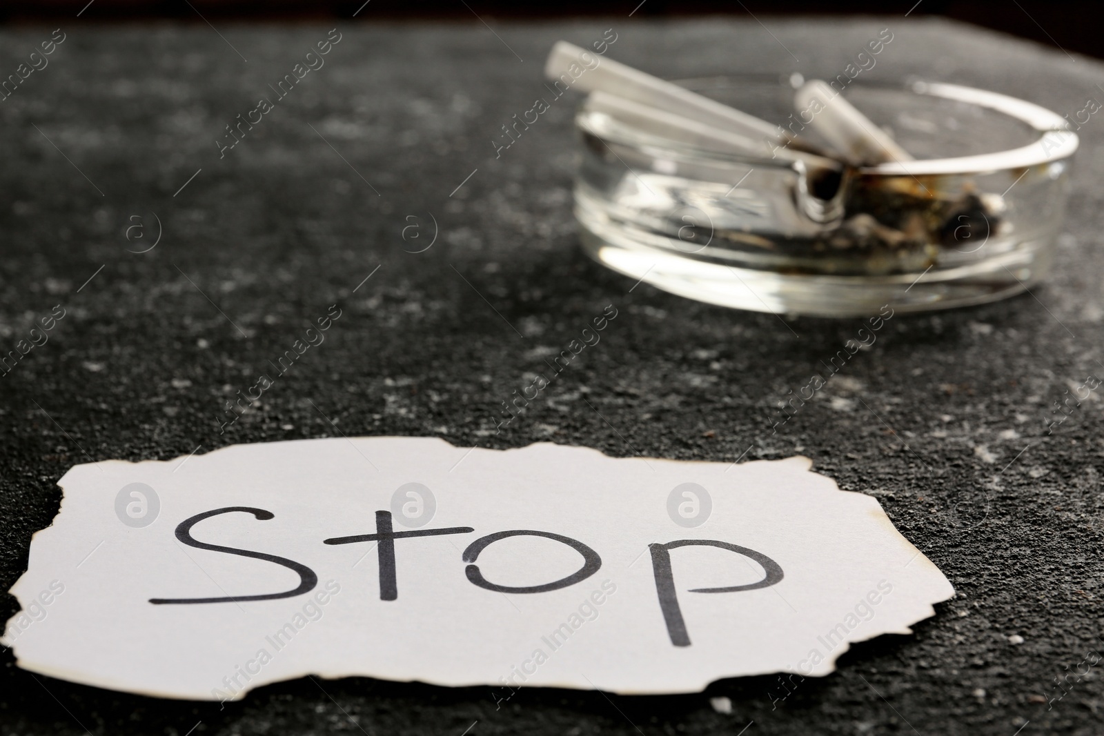 Photo of Ashtray with burnt cigarettes and word Stop written on paper on black textured table, closeup. No smoking concept