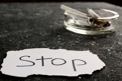 Ashtray with burnt cigarettes and word Stop written on paper on black textured table, closeup. No smoking concept