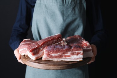 Photo of Woman holding wooden board with pieces of raw pork belly on black background, closeup