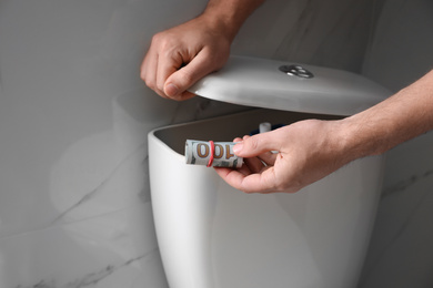 Photo of Man hiding money in toilet tank indoors, closeup. Financial savings