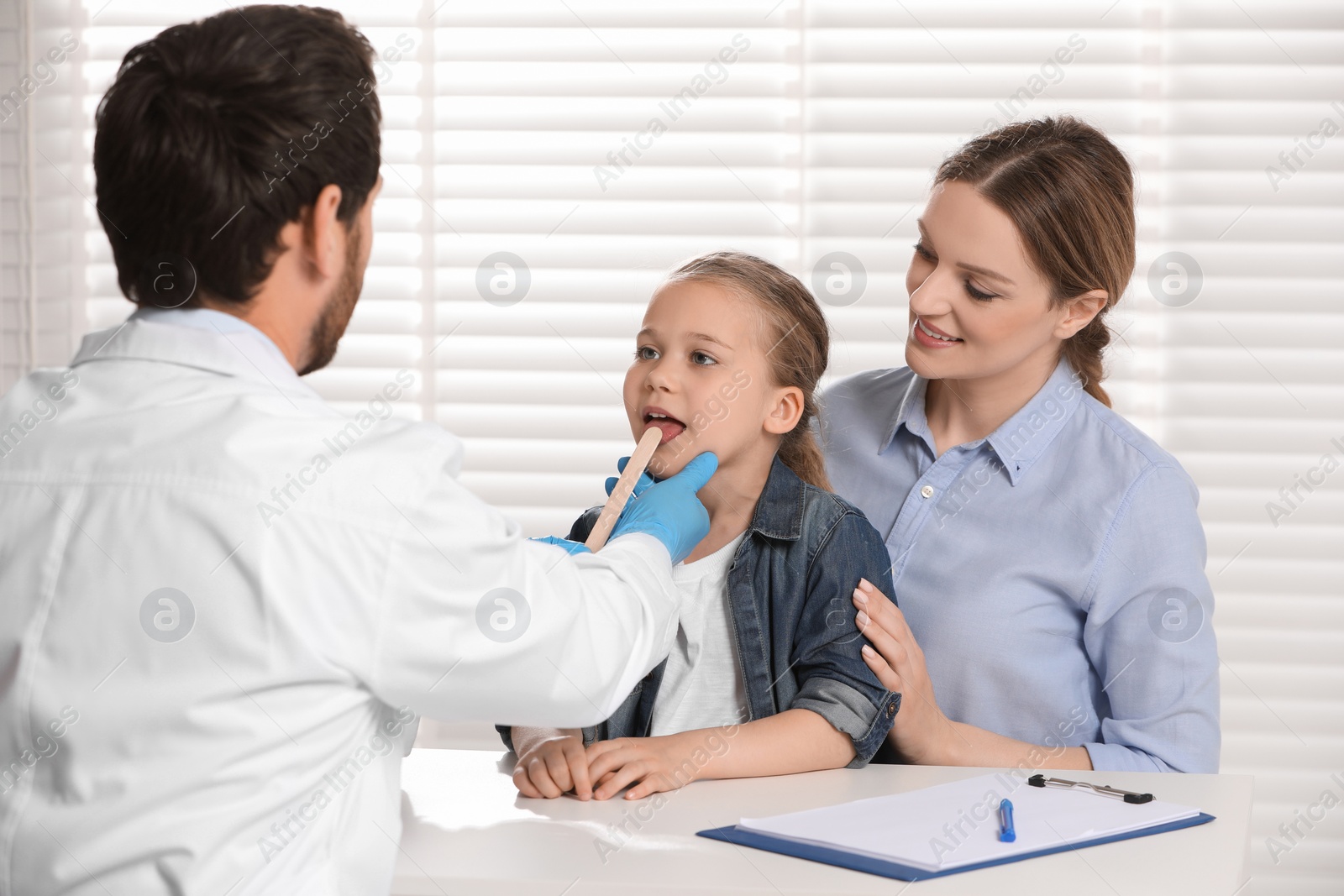 Photo of Doctor examining girl`s oral cavity with tongue depressor near her mother indoors