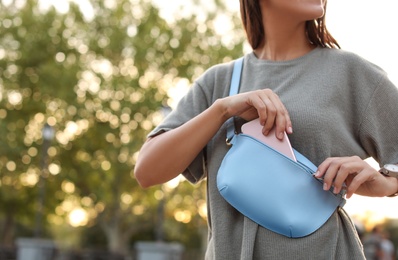 Young woman putting smartphone in stylish bum bag outdoors, closeup