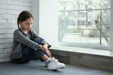 Photo of Sad little girl sitting on floor near window indoors, space for text
