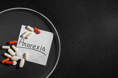 Photo of Note paper with word Anorexia, plate and pills on black background, flat lay. Space for text