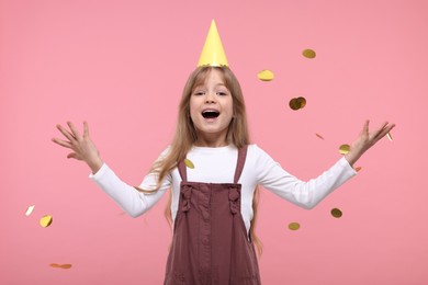 Happy little girl in party hat throwing confetti on pink background