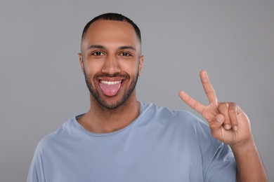 Photo of Happy young man showing his tongue and V-sign on light grey background