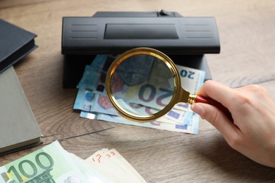 Photo of Woman checking Euro banknotes with currency detector and magnifying glass at wooden table, closeup. Money examination device