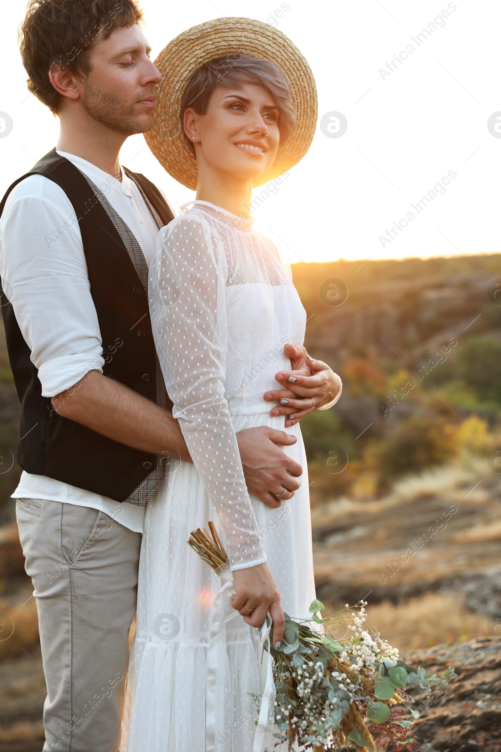 Photo of Happy newlyweds with beautiful field bouquet standing on rock at sunset