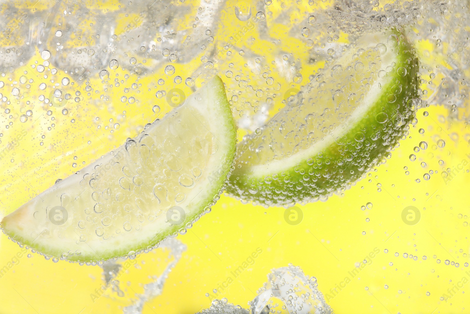 Photo of Juicy lime slices and ice cubes in soda water against yellow background, closeup