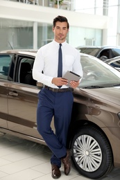 Young salesman with tablet near car in modern dealership