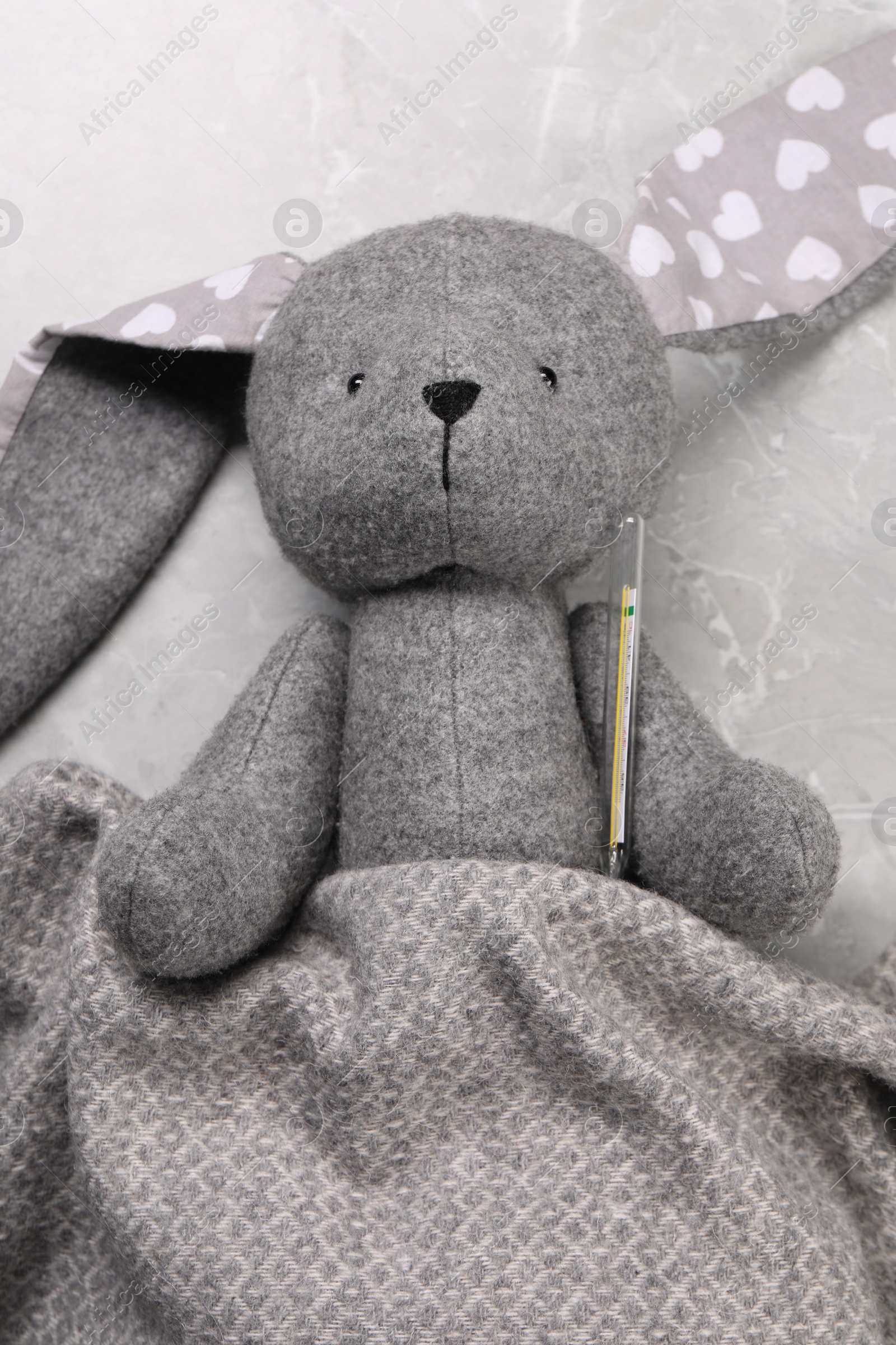 Photo of Toy bunny with thermometer under blanket on gray marble background, top view