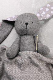 Photo of Toy bunny with thermometer under blanket on gray marble background, top view