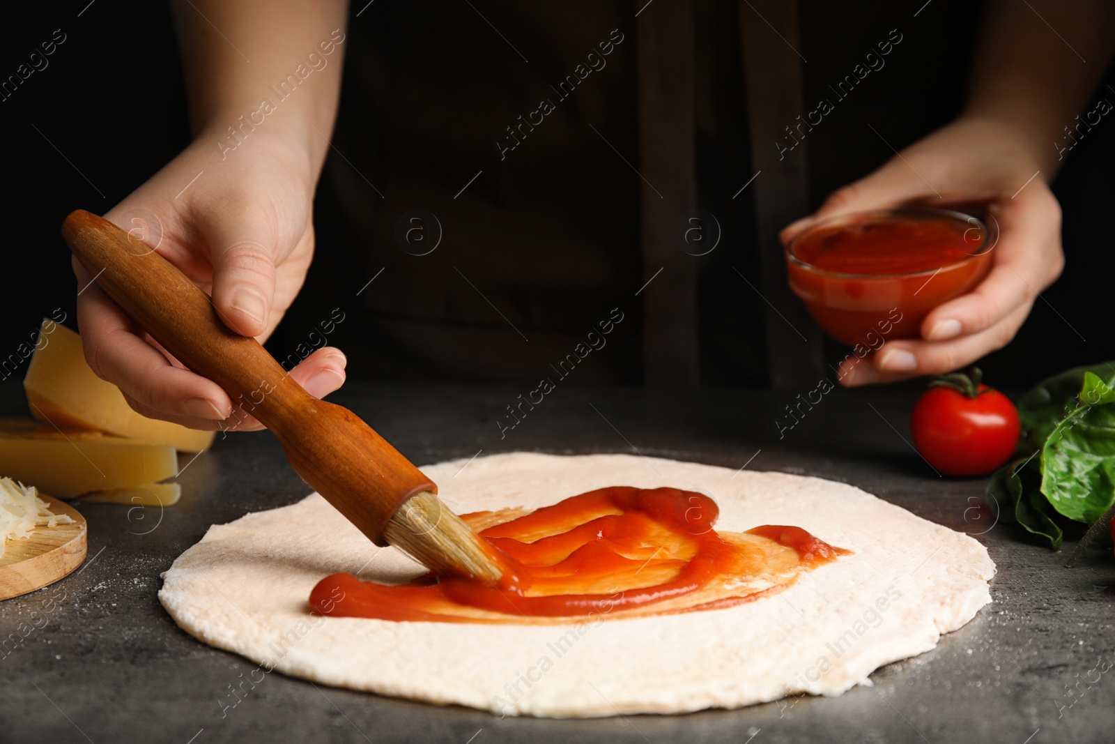 Photo of Woman spreading tomato sauce onto pizza crust at grey table, closeup