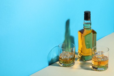 Photo of Whiskey with ice cubes in glasses and bottle on white table against light blue background, space for text