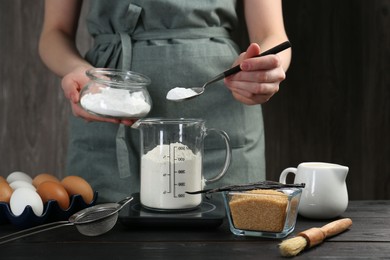 Woman adding baking powder into measuring cup at black wooden table, closeup