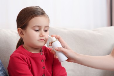 Mother using nasal spray to treat her little daughter on sofa indoors, closeup