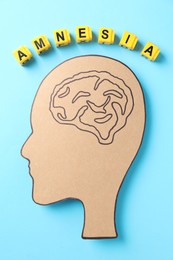 Photo of Yellow cubes with word Amnesia and human head cutout with drawing of brain on light blue background, top view