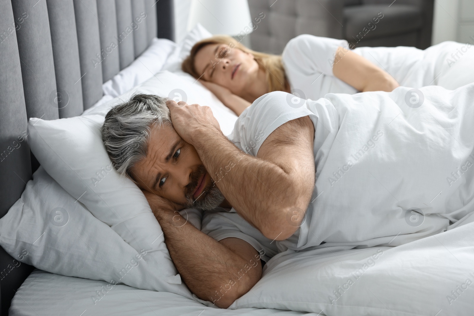 Photo of Irritated man covering his ears in bed at home Problem with snoring wife