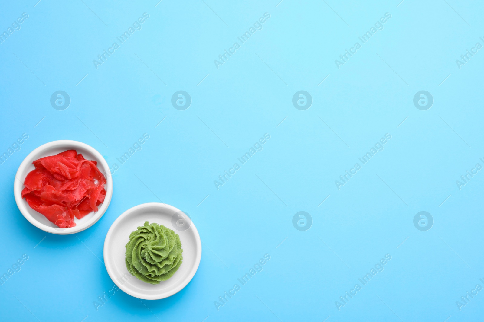 Photo of Bowls with swirl of wasabi paste and pickled ginger on light blue background, flat lay. Space for text