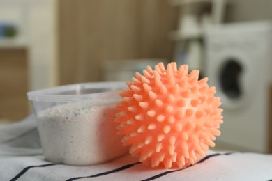 Photo of Orange dryer ball and detergent on clean clothing in laundry room, closeup