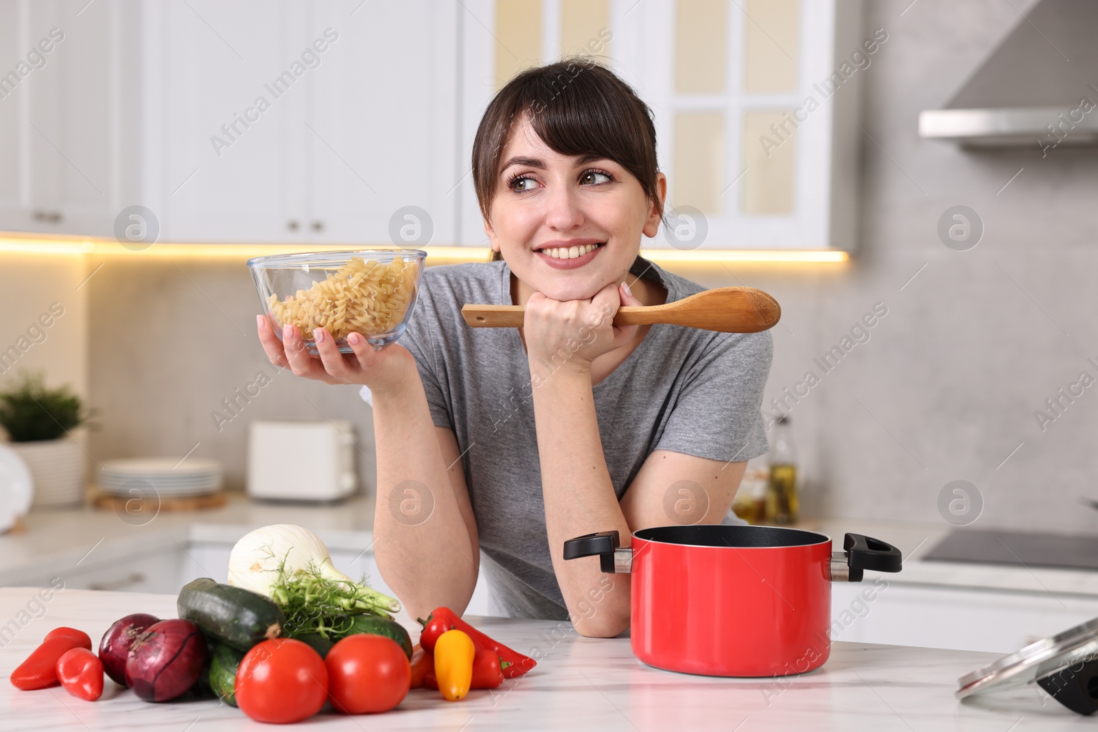 Photo of Happy young housewife with spoon and raw pasta at white marble table in kitchen. Cooking process