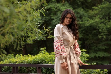 Photo of Beautiful woman wearing embroidered dress near wooden railing in countryside. Ukrainian national clothes