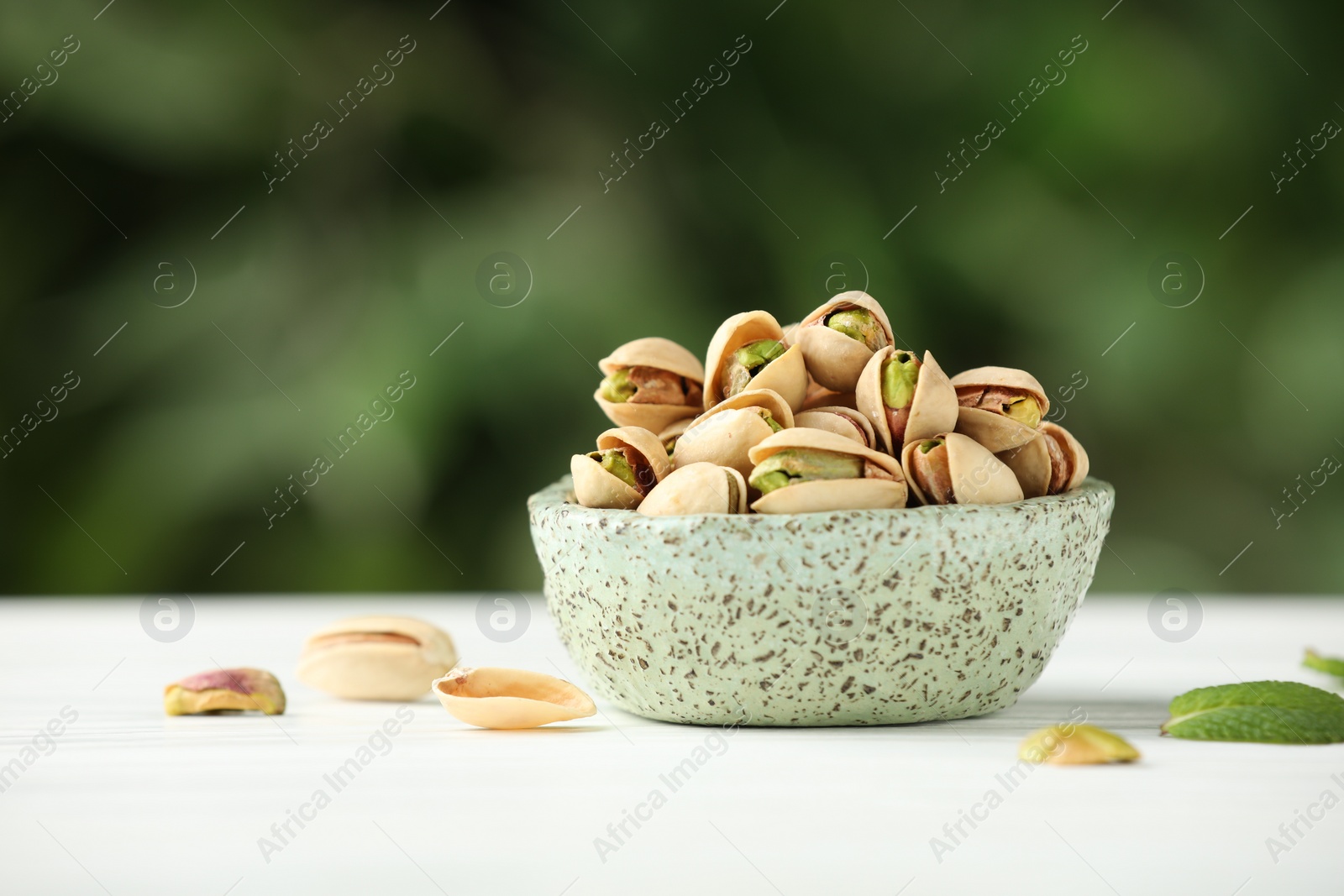 Photo of Tasty pistachios in bowl on white table against blurred background. Space for text