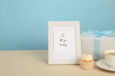 Happy Father's Day. Tasty cupcake, cup, card with phrase I Love You, Daddy in frame and gift box on beige table, closeup. Space for text