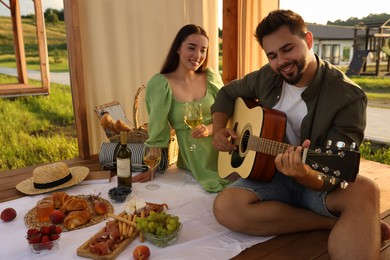 Photo of Romantic date. Beautiful woman with glass of wine and her boyfriend playing guitar during picnic in wooden gazebo