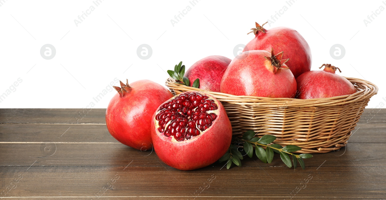 Photo of Fresh pomegranates in wicker basket and green leaves on wooden table against white background