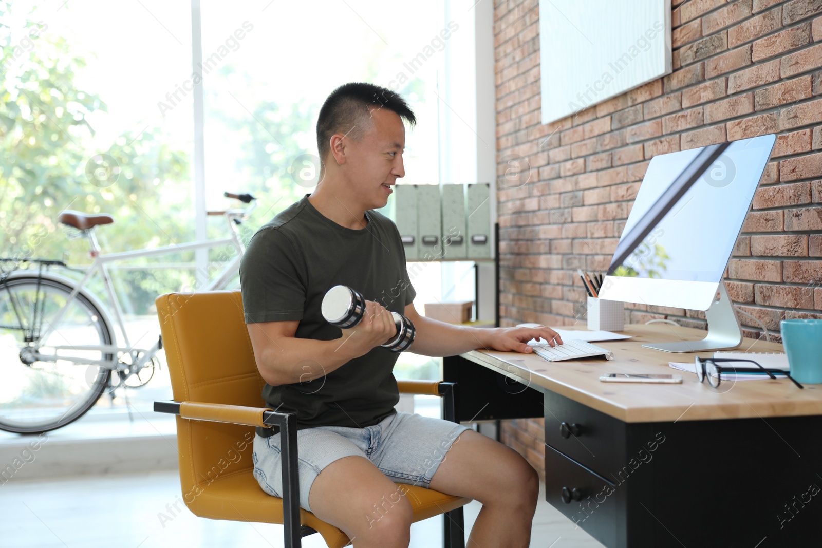 Photo of Young man lifting weights and using computer in office. Workplace fitness