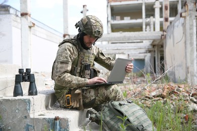 Military mission. Soldier in uniform using laptop near abandoned building outside