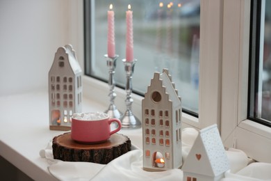 Photo of Beautiful house shaped candle holders and hot dink with marshmallow on windowsill indoors