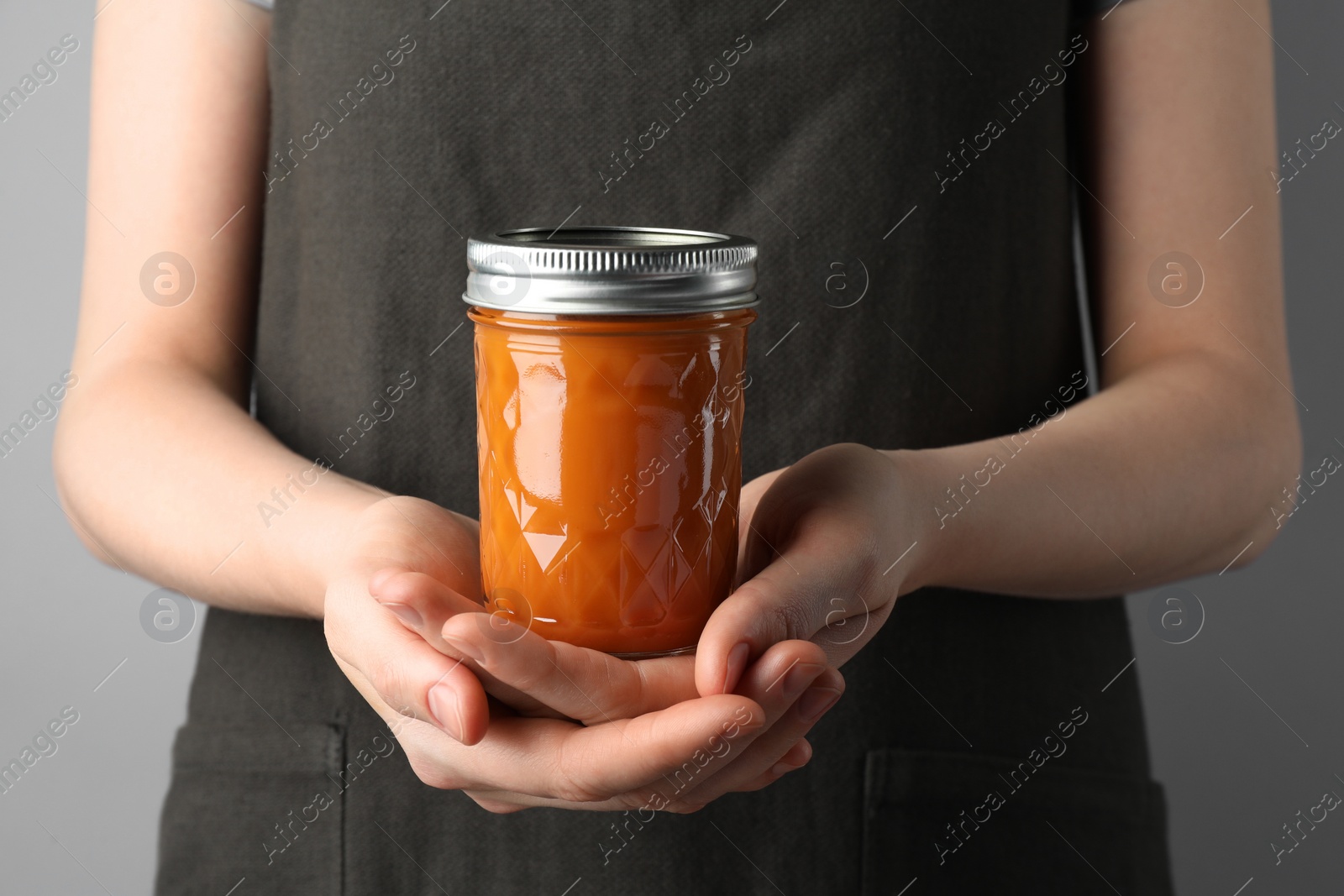 Photo of Woman holding glass jar of delicious persimmon jam on gray background, closeup