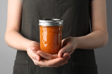 Photo of Woman holding glass jar of delicious persimmon jam on gray background, closeup